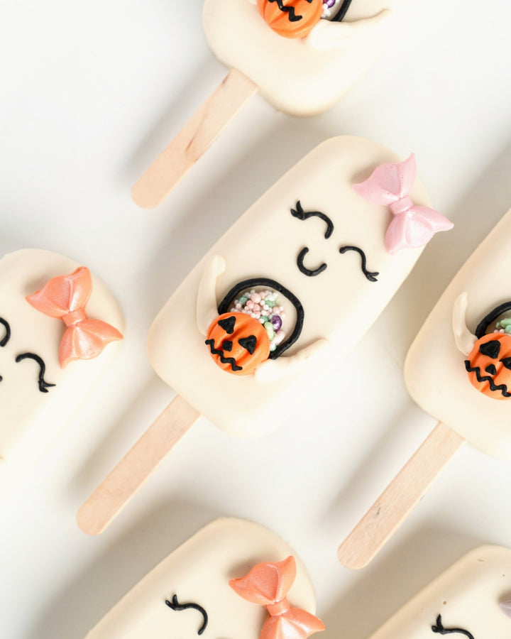 Ghost cakesicles with pastel bows holding jack-o'-lanterns