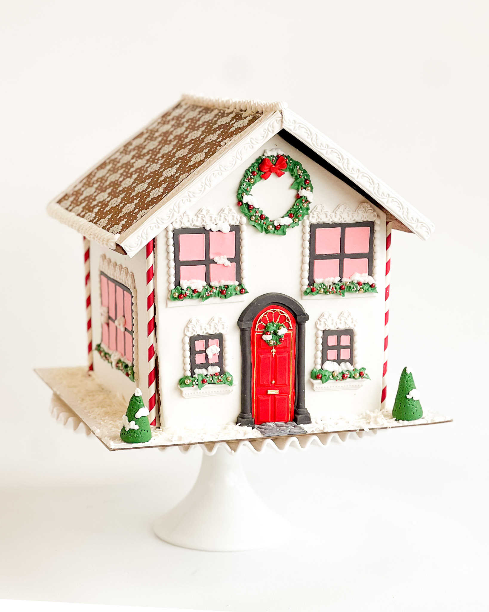 Classic holiday gingerbread house with a red door, candy cane accents, snow-dusted roof, and festive wreath, placed on a white cake stand