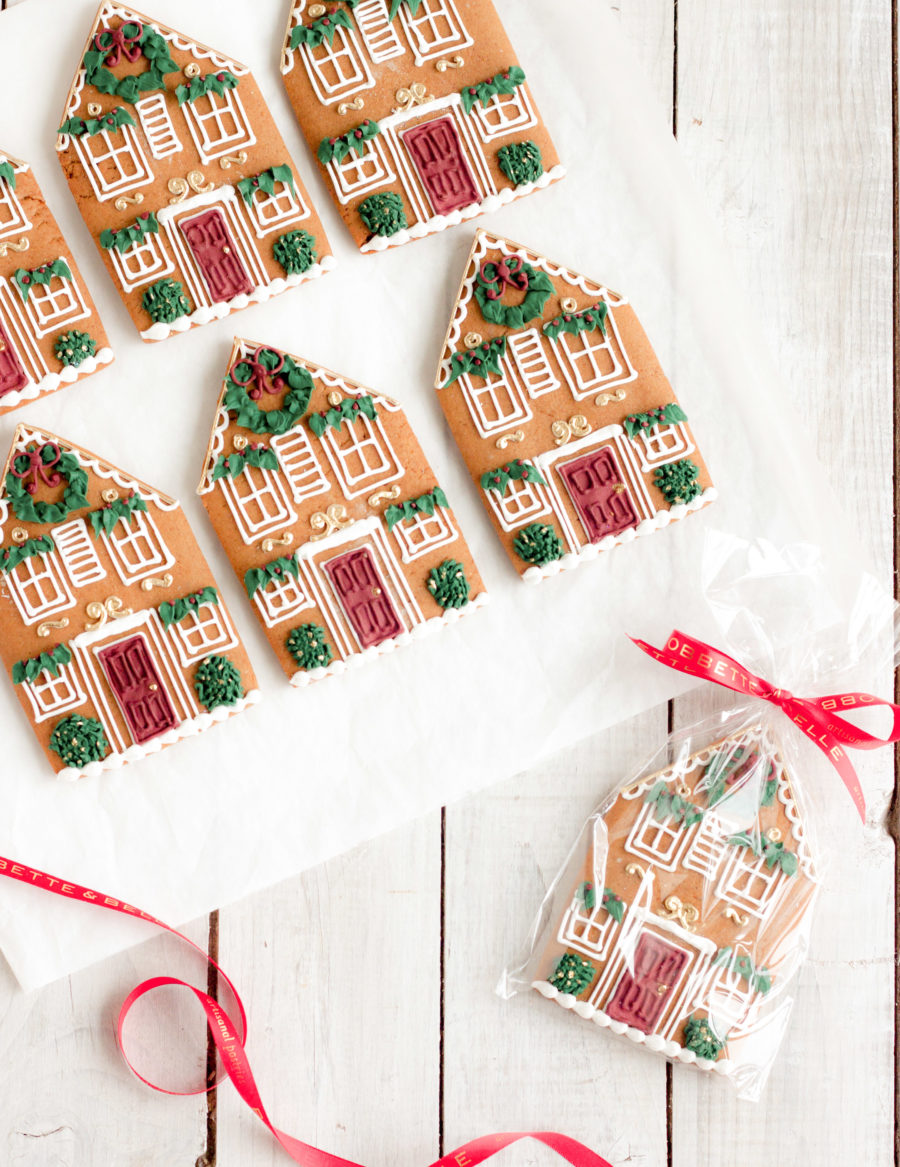 Victorian gingerbread house cookies featuring detailed royal icing designs, red doors, wreaths, and greenery, individually wrapped with ribbons.