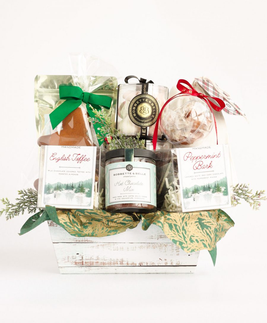 A holiday-themed gift basket filled with festive treats, including chocolate cookies, caramel corn, English toffee, peppermint bark, hot chocolate mix, vanilla marshmallows, holiday meringues, a gingerbread cookie, and salted caramels in a bauble.