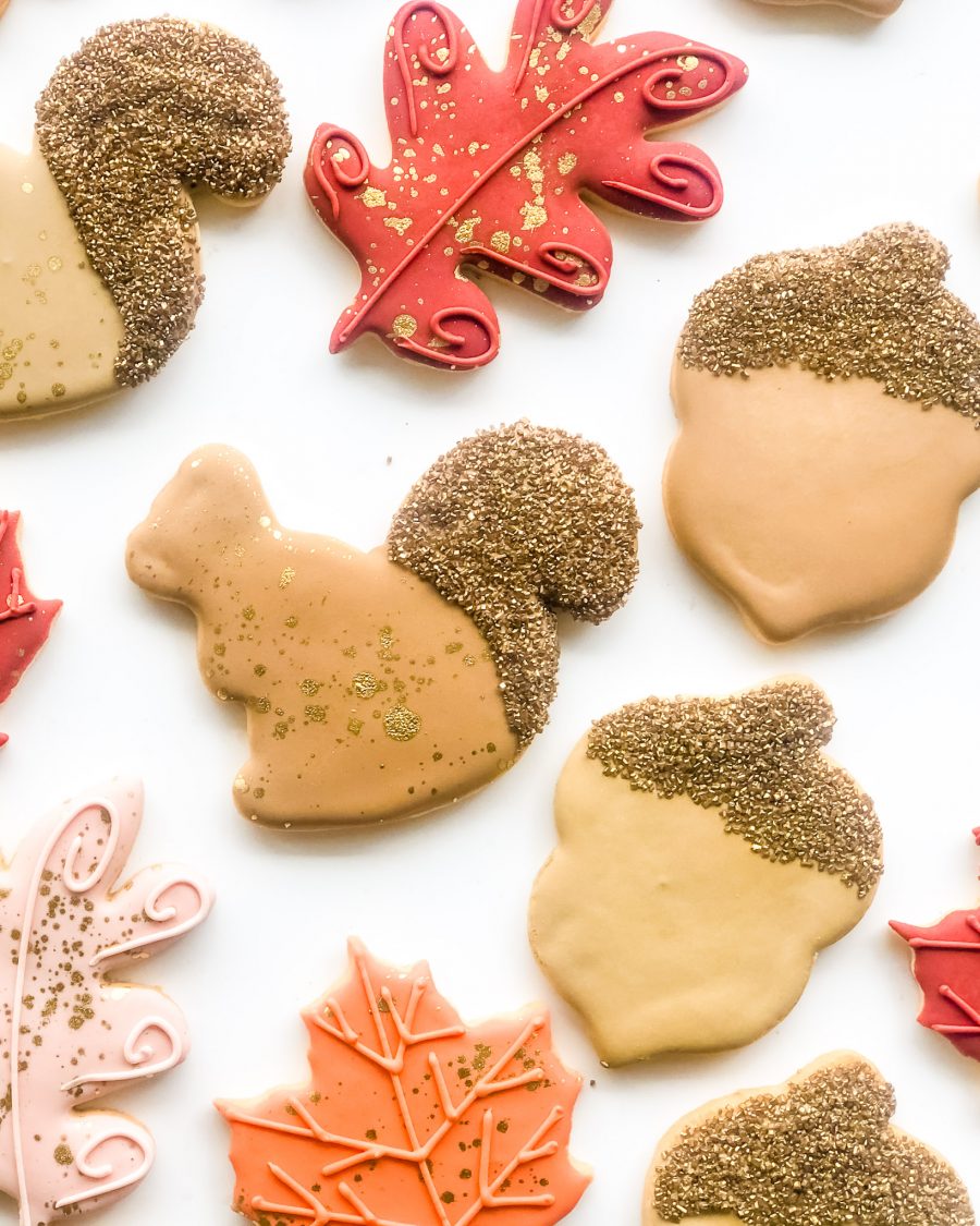 Thanksgiving-themed sugar cookies shaped like oak and maple leaves in fall colours, with a squirrel and glittering acorn, packaged in clear bags with ribbon ties