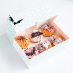 Close-up of a 6-pack of Halloween cupcakes with pastel buttercream swirls, featuring fondant ghosts, bats, and 'Happy Halloween' toppers.