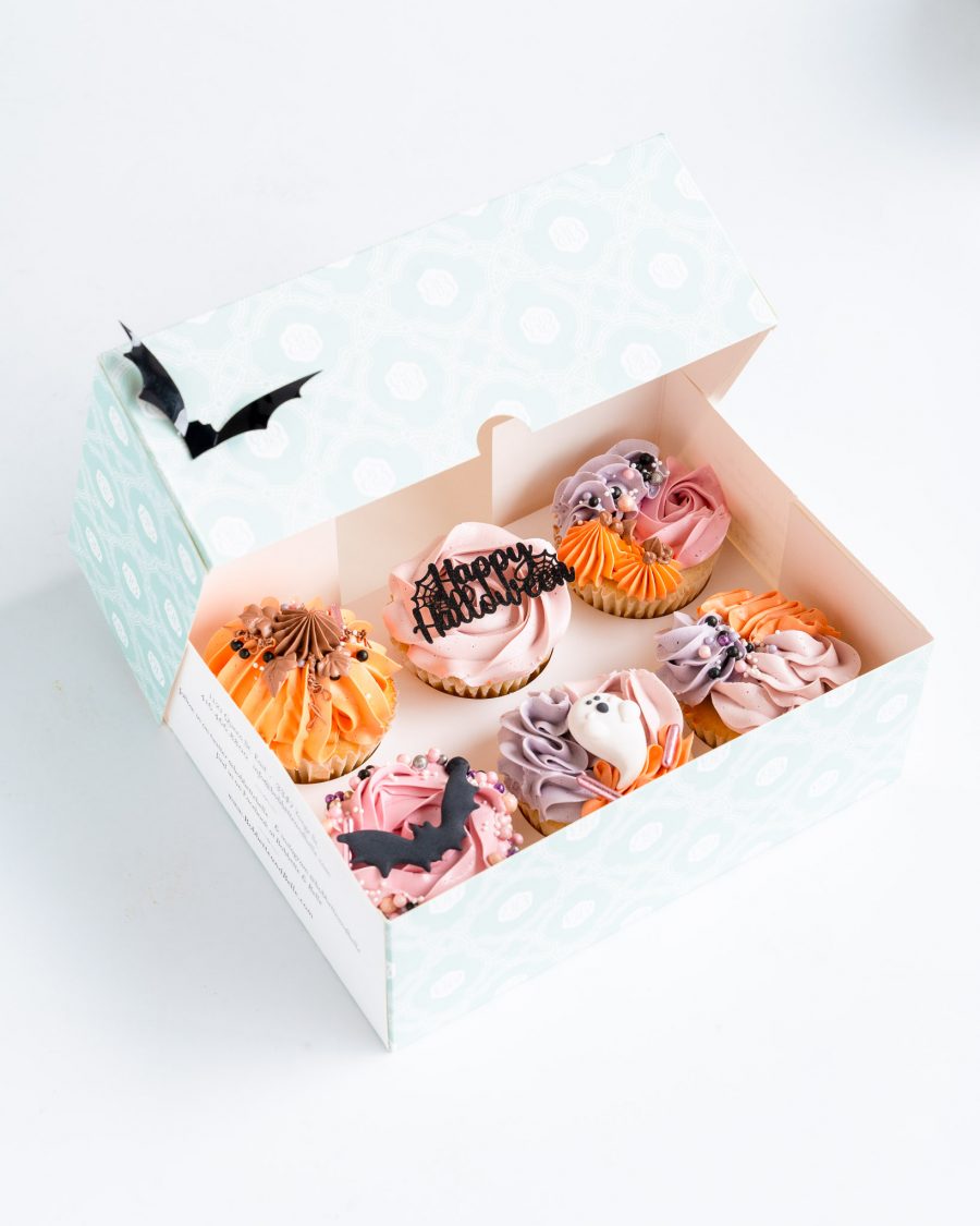 Close-up of a 6-pack of Halloween cupcakes with pastel buttercream swirls, featuring fondant ghosts, bats, and 'Happy Halloween' toppers.