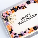 A rectangular sheet cake decorated for Halloween, with a smooth white icing base and a colorful buttercream border. The border features intricate piping in shades of orange, black, purple, and pink, forming a festive and playful design. On one corner, a gold spider web is piped delicately, complete with a small decorative spider. The center of the cake displays the words "Happy Halloween" in a bold, elegant font, creating a clean and stylish look. The cake sits on a silver cake board, making it a beautiful centerpiece for any Halloween gathering.