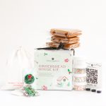 A gingerbread house kit featuring pre-baked gingerbread pieces, royal icing mix, food colors, piping bags, sugar decorations, and a clear branded box with ribbon.