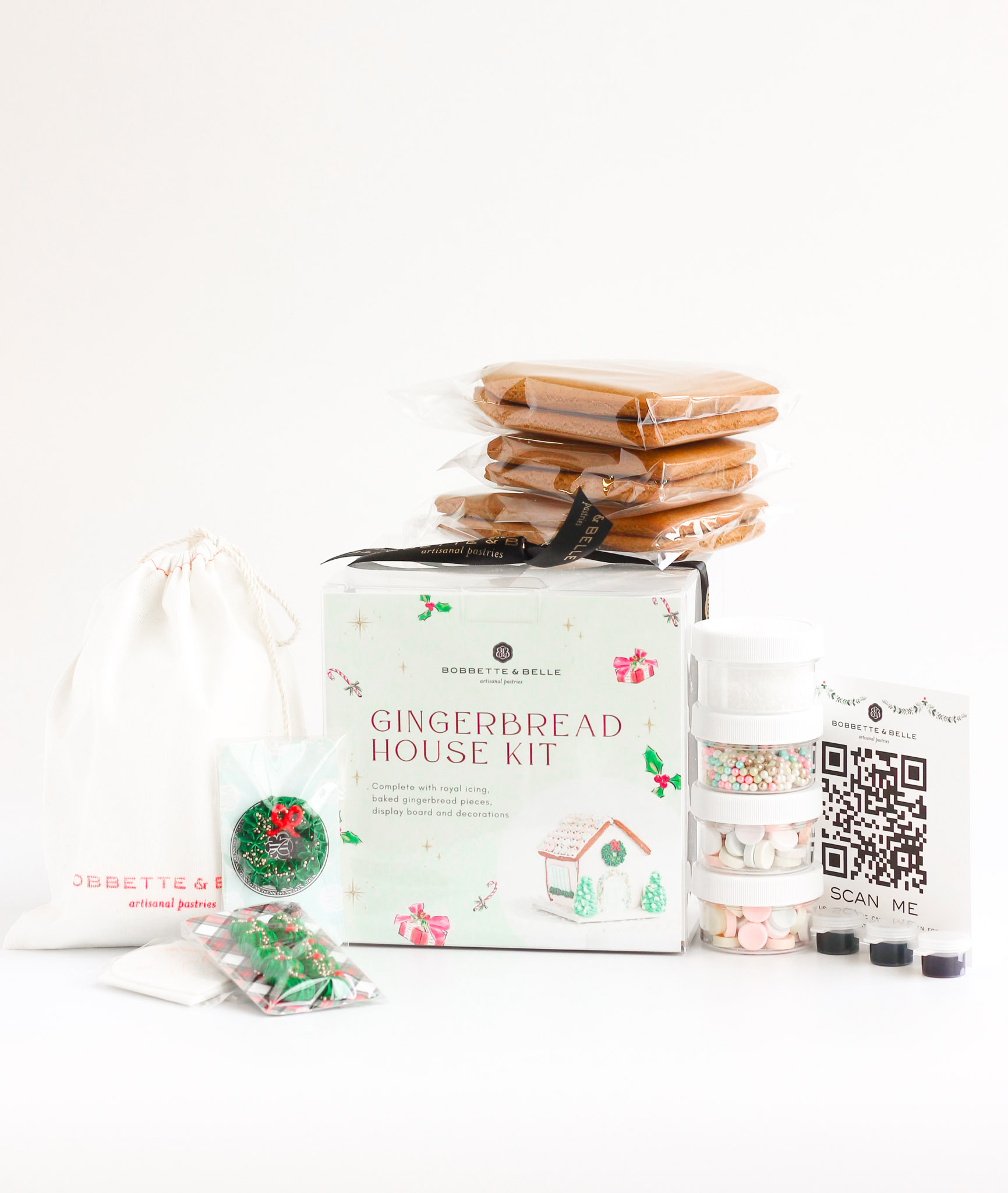 A gingerbread house kit featuring pre-baked gingerbread pieces, royal icing mix, food colors, piping bags, sugar decorations, and a clear branded box with ribbon.