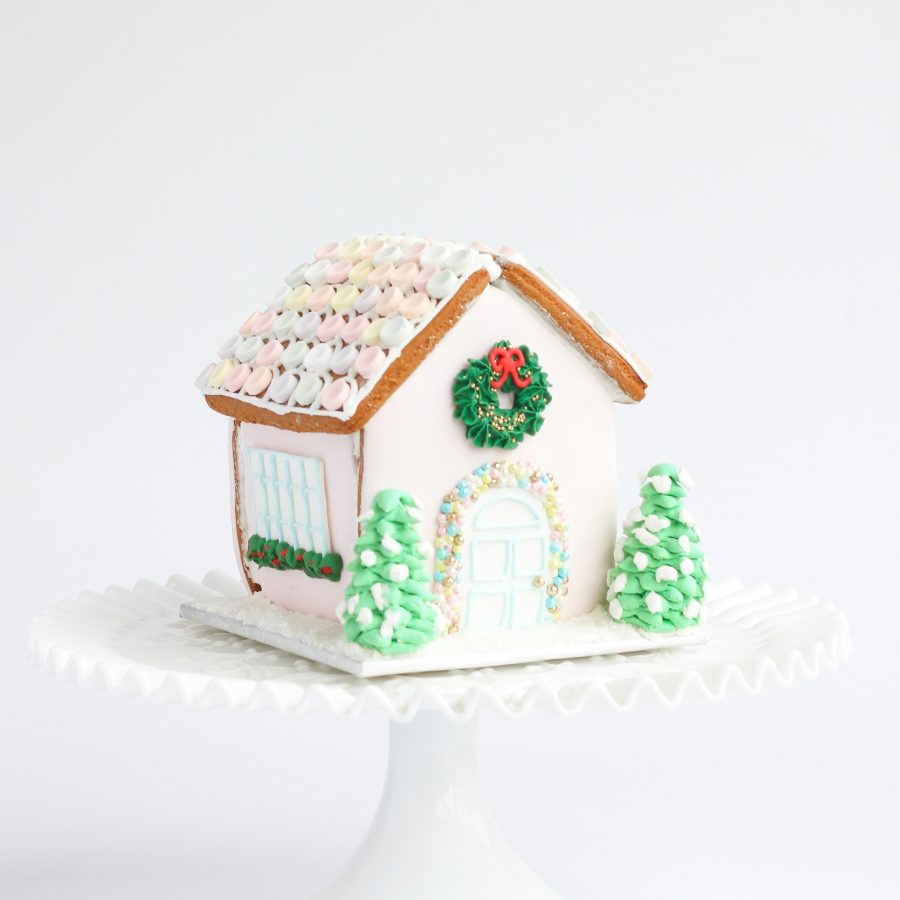 A fully assembled gingerbread house decorated with royal icing, sugar dragees, a piped wreath, and sparkling sugar snow, sitting on a festive board.