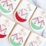 Hand-decorated festive ornament sugar cookies with red, green, and gold accents, shaped like Christmas ornaments, displayed on a marble surface.