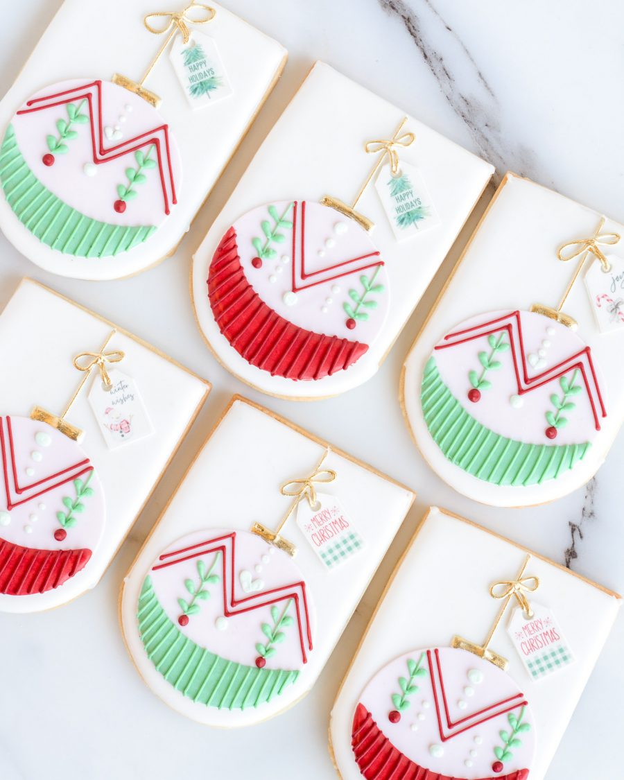 Hand-decorated festive ornament sugar cookies with red, green, and gold accents, shaped like Christmas ornaments, displayed on a marble surface.