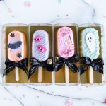 The image shows four Halloween-themed cakecicles laid out side by side on a marble surface. Each cakecicle has a distinct design: the first is coated in a pastel peach color with a black bat and star details, the second is a mint green cakecicle with a cute white ghost, the third is purple with pink vampire lips, and the fourth is pink with colorful sprinkles and the word "BOO!" written in playful, bright letters. The cakecicles are decorated with drizzles and embellishments, creating a fun, festive, and whimsical presentation perfect for Halloween.