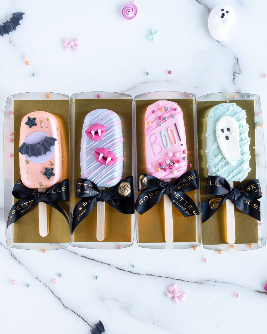 The image shows four Halloween-themed cakecicles laid out side by side on a marble surface. Each cakecicle has a distinct design: the first is coated in a pastel peach color with a black bat and star details, the second is a mint green cakecicle with a cute white ghost, the third is purple with pink vampire lips, and the fourth is pink with colorful sprinkles and the word "BOO!" written in playful, bright letters. The cakecicles are decorated with drizzles and embellishments, creating a fun, festive, and whimsical presentation perfect for Halloween.