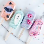 The image shows four Halloween-Themed Cake Pops laid out side by side on a marble surface. Each cakecicle has a distinct design: the first is coated in a pastel peach color with a black bat and star details, the second is a mint green cakecicle with a cute white ghost, the third is purple with pink vampire lips, and the fourth is pink with colorful sprinkles and the word "BOO!" written in playful, bright letters. The cakecicles are decorated with drizzles and embellishments, creating a fun, festive, and whimsical presentation perfect for Halloween.