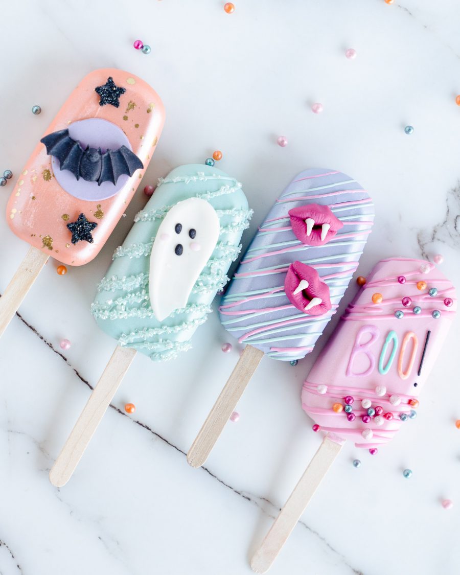 The image shows four Halloween-Themed Cake Pops laid out side by side on a marble surface. Each cakecicle has a distinct design: the first is coated in a pastel peach color with a black bat and star details, the second is a mint green cakecicle with a cute white ghost, the third is purple with pink vampire lips, and the fourth is pink with colorful sprinkles and the word "BOO!" written in playful, bright letters. The cakecicles are decorated with drizzles and embellishments, creating a fun, festive, and whimsical presentation perfect for Halloween.