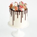 A tall Festive Christmas Cake decorated with chocolate ganache drips, gingerbread men, and candy canes on a white cake stand, perfect for holiday celebrations.