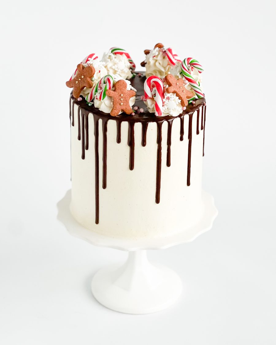 A tall Festive Christmas Cake decorated with chocolate ganache drips, gingerbread men, and candy canes on a white cake stand, perfect for holiday celebrations.