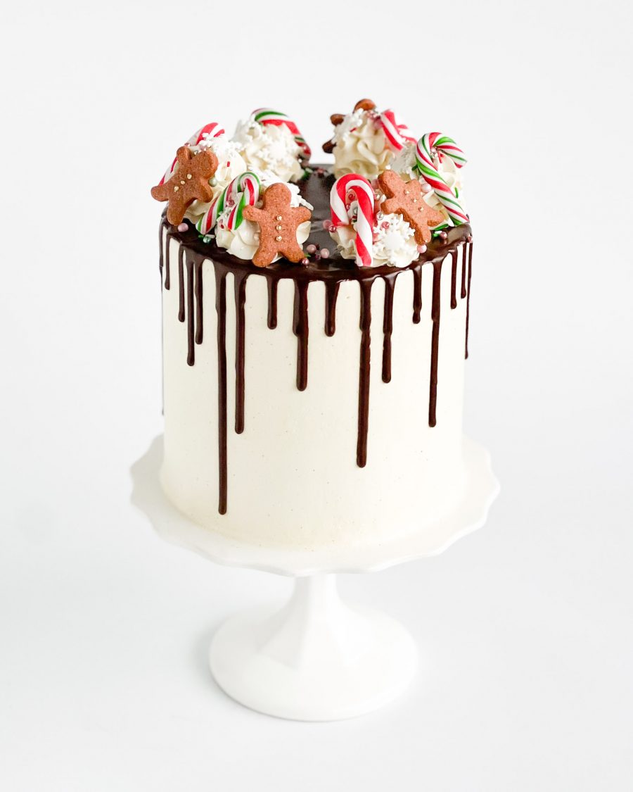 Festive holiday drip cake topped with gingerbread cookies, candy canes, whipped frosting swirls, and chocolate ganache drips.