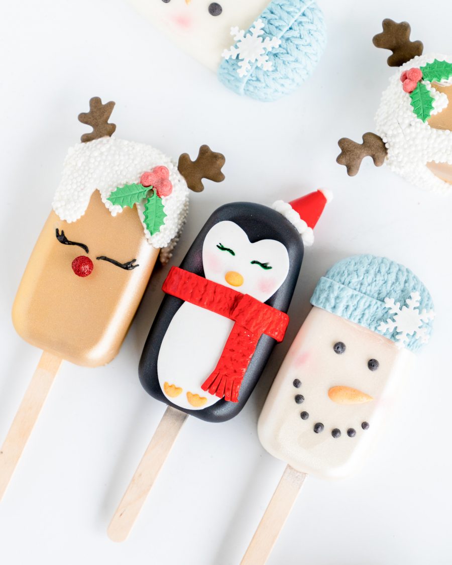 Holiday-themed cake popsicles featuring hand-decorated designs of a reindeer with holly accents, a penguin with a red scarf and Santa hat, and a snowman with a blue knitted hat and snowflake decoration.