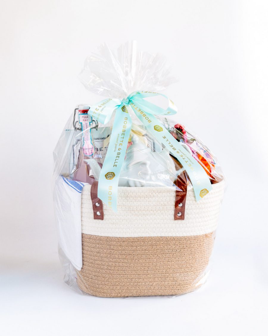 A cream and natural brown woven tote bag filled with Mother's Day treats including a wildflower bouquet, Bobbette & Belle recipe book, sparkling lemonade, decorative pastel vanilla cupcake, fresh blueberry scones, Earl Grey tea and a china teapot