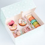 A box filled with Vanilla Rose swirl cupcakes with Happy Mother's Day heart shaped plaque, sugar butterfly and decorative buttercream piping in shades of soft pink, blue lavender and turquoise. Packaged with 6 colour coordinated french macarons