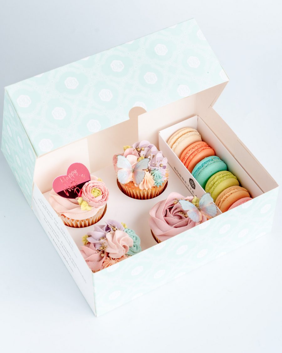 A box filled with Vanilla Rose swirl cupcakes with Happy Mother's Day heart shaped plaque, sugar butterfly and decorative buttercream piping in shades of soft pink, blue lavender and turquoise. Packaged with 6 colour coordinated french macarons