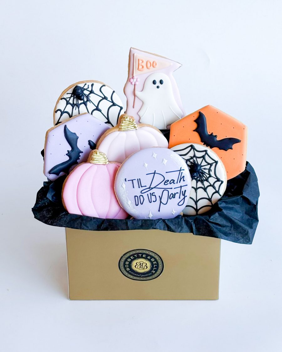 A Halloween-themed gift box filled with an assortment of hand-decorated sugar cookies. The box includes cookies shaped like ghosts holding "Boo" flags, pastel pumpkins with golden tops, spiderweb designs, orange hexagons with black bats, and purple crystal balls with the phrase "'Til Death Do Us Party." Each cookie is intricately decorated, showcasing vibrant colors and festive themes, arranged beautifully in a gift box lined with black tissue paper, creating a delightful Halloween presentation.