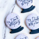 A set of round, pastel purple cookies styled as crystal balls, each displaying the phrase "'Til Death Do Us Party" in stylish black script. The cookies have a dark base and small white stars, arranged on a marble background with matching icing bags nearby, creating a chic, spooky vibe.