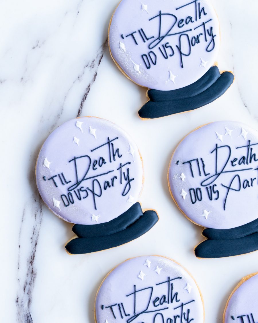 A set of round, pastel purple cookies styled as crystal balls, each displaying the phrase "'Til Death Do Us Party" in stylish black script. The cookies have a dark base and small white stars, arranged on a marble background with matching icing bags nearby, creating a chic, spooky vibe.