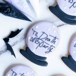 An assortment of round sugar cookies designed to look like crystal balls, with pastel purple icing and a smooth black base. The message "'Til Death Do Us Party" is piped in black, surrounded by small white star accents for a gothic and elegant touch. The cookies are beautifully arranged on a marble surface, accompanied by black and purple icing bags and bat-shaped accents, creating a festive and stylish Halloween display.