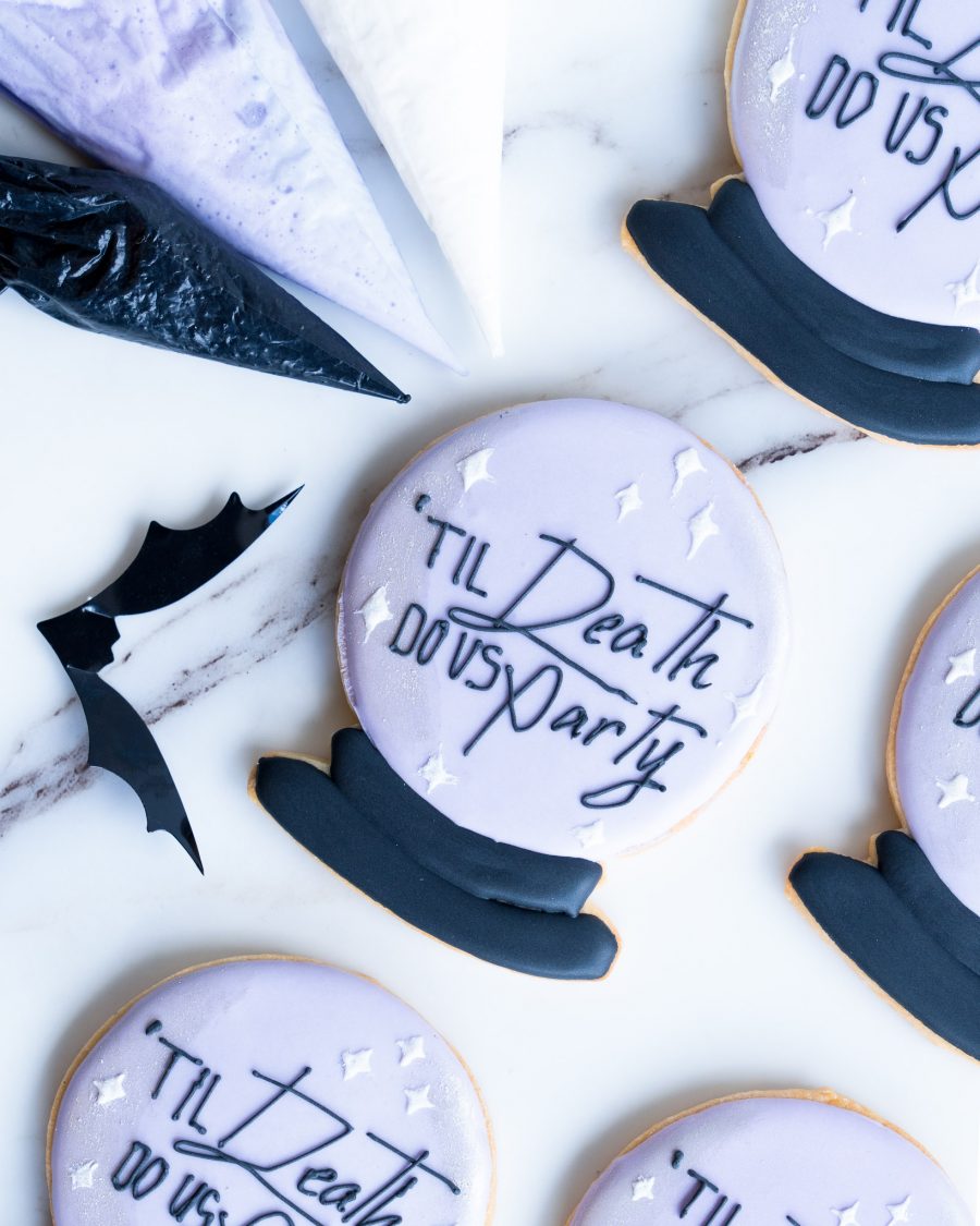 An assortment of round sugar cookies designed to look like crystal balls, with pastel purple icing and a smooth black base. The message "'Til Death Do Us Party" is piped in black, surrounded by small white star accents for a gothic and elegant touch. The cookies are beautifully arranged on a marble surface, accompanied by black and purple icing bags and bat-shaped accents, creating a festive and stylish Halloween display.