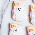 A close-up view of hand-decorated ghost-shaped sugar cookies displayed on a marble surface. The cookies feature a soft, pastel pink and white color palette, with a playful ghost design. Each ghost holds a pastel pink flag with the word "Boo" written in orange icing, accented by a small pink flower. The cookies have smooth, carefully piped icing details that make them both visually appealing and delicious. These Halloween treats are perfect for adding a fun, festive touch to any celebration.