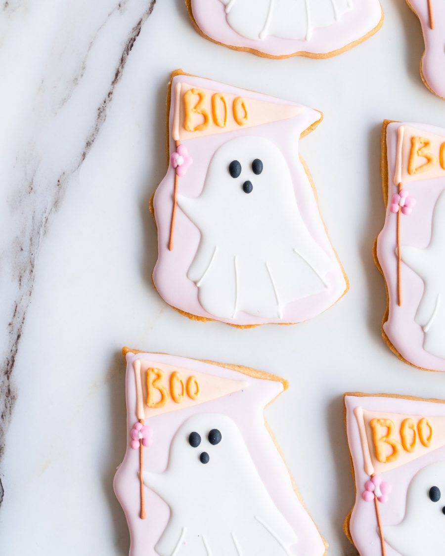 A close-up view of hand-decorated ghost-shaped sugar cookies displayed on a marble surface. The cookies feature a soft, pastel pink and white color palette, with a playful ghost design. Each ghost holds a pastel pink flag with the word "Boo" written in orange icing, accented by a small pink flower. The cookies have smooth, carefully piped icing details that make them both visually appealing and delicious. These Halloween treats are perfect for adding a fun, festive touch to any celebration.