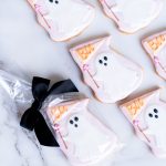 An assortment of hand-decorated ghost sugar cookies arranged on a marble surface. The cookies feature a friendly ghost design with pastel pink and white icing, holding a "Boo" flag adorned with a tiny pink flower. One cookie is individually wrapped in a clear bag and tied with a black satin bow, making it perfect for gifting or as a stylish party favor. The cookies showcase delicate piping details, providing a soft, buttery texture and a festive appearance that captures the spirit of Halloween.