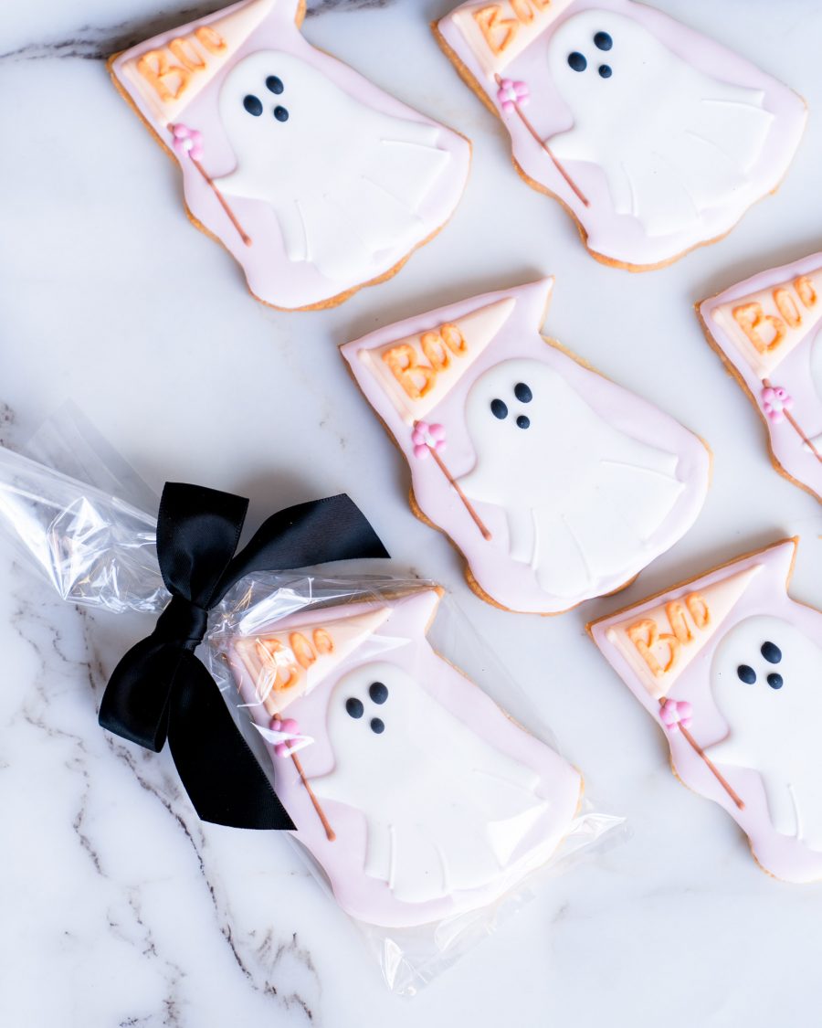 An assortment of hand-decorated ghost sugar cookies arranged on a marble surface. The cookies feature a friendly ghost design with pastel pink and white icing, holding a "Boo" flag adorned with a tiny pink flower. One cookie is individually wrapped in a clear bag and tied with a black satin bow, making it perfect for gifting or as a stylish party favor. The cookies showcase delicate piping details, providing a soft, buttery texture and a festive appearance that captures the spirit of Halloween.