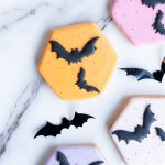 A collection of hand-decorated sugar cookies laid out on a marble surface. Each cookie is shaped as a hexagon and decorated with pastel-colored icing in shades of orange, pink, and purple. Smooth black bat designs are intricately placed on each cookie, creating a contrast that’s both elegant and spooky. The vibrant yet soft colors make these treats a festive addition to Halloween celebrations, ideal for parties, gift boxes, or as a fun seasonal snack.