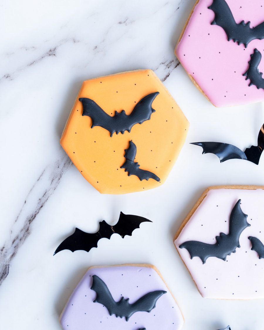 A collection of hand-decorated sugar cookies laid out on a marble surface. Each cookie is shaped as a hexagon and decorated with pastel-colored icing in shades of orange, pink, and purple. Smooth black bat designs are intricately placed on each cookie, creating a contrast that’s both elegant and spooky. The vibrant yet soft colors make these treats a festive addition to Halloween celebrations, ideal for parties, gift boxes, or as a fun seasonal snack.