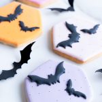 A selection of hexagon-shaped sugar cookies with pastel orange, pink, and purple icing, each adorned with smooth, black bat designs. Displayed on a marble background, these hand-decorated cookies bring a mix of festive color and spooky charm, perfect for Halloween events and gifts.