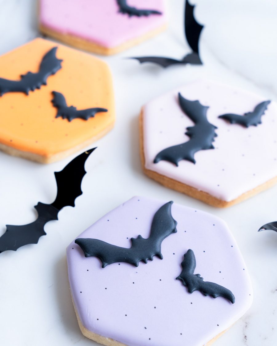 A selection of hexagon-shaped sugar cookies with pastel orange, pink, and purple icing, each adorned with smooth, black bat designs. Displayed on a marble background, these hand-decorated cookies bring a mix of festive color and spooky charm, perfect for Halloween events and gifts.