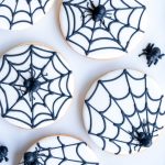 A collection of round sugar cookies, each decorated with a smooth white icing base and a detailed black spiderweb design. Small, edible spiders are placed atop the web, adding a spooky yet cute touch to the cookies. Arranged on a white surface, these hand-decorated treats showcase clean lines and precise piping, making them a delightful and festive addition to any Halloween celebration.