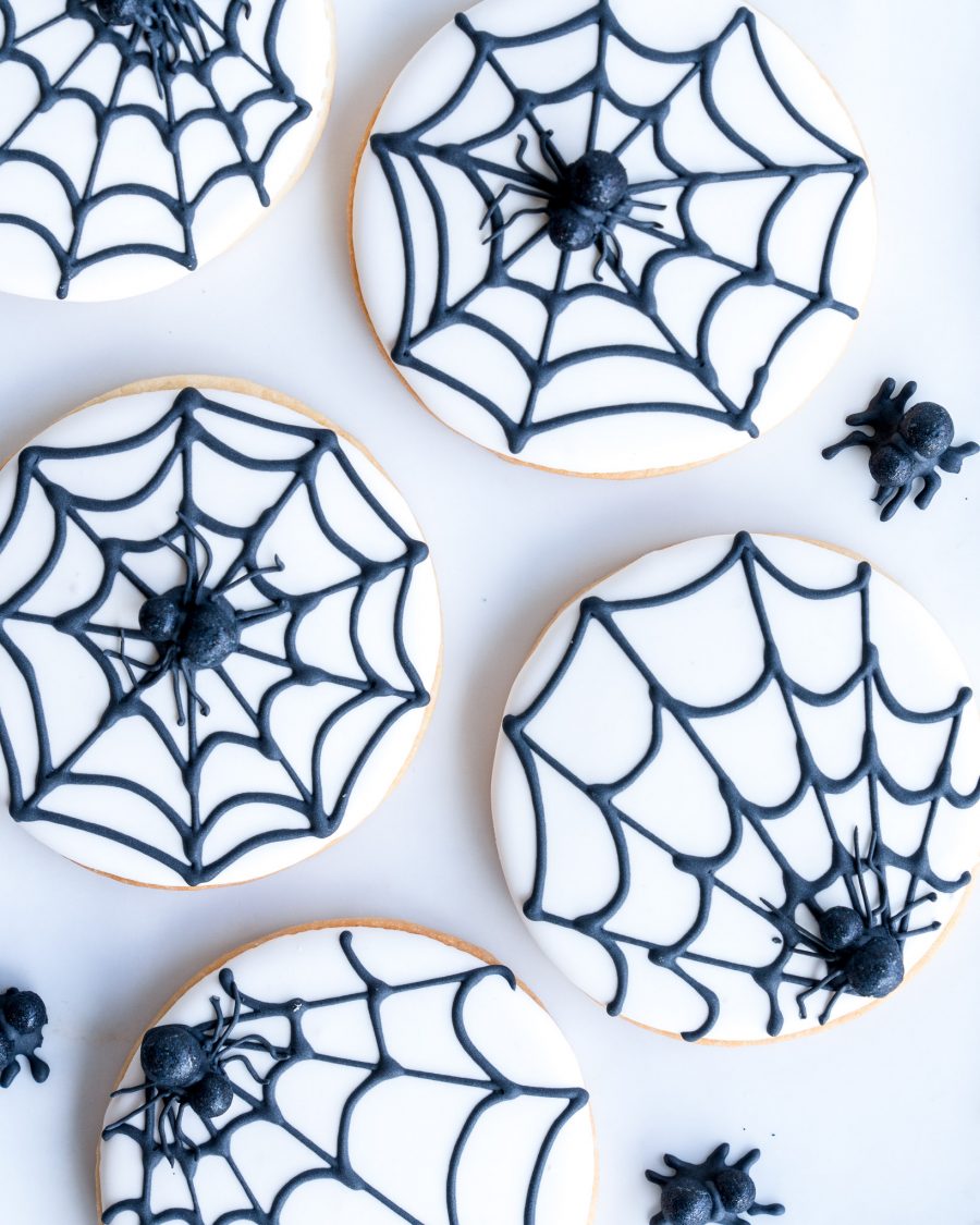 A collection of round sugar cookies, each decorated with a smooth white icing base and a detailed black spiderweb design. Small, edible spiders are placed atop the web, adding a spooky yet cute touch to the cookies. Arranged on a white surface, these hand-decorated treats showcase clean lines and precise piping, making them a delightful and festive addition to any Halloween celebration.