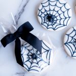 A display of round sugar cookies, each featuring a smooth white icing base with finely piped black spiderwebs. Atop each web sits a small, edible spider, adding a playful yet eerie element. The cookies are neatly arranged on a white surface, showcasing their detailed design, making them an ideal Halloween treat for parties or gifts.