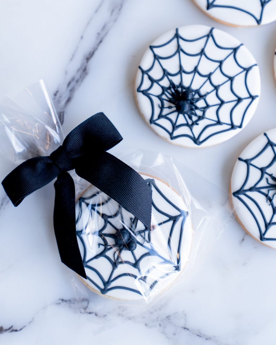 A display of round sugar cookies, each featuring a smooth white icing base with finely piped black spiderwebs. Atop each web sits a small, edible spider, adding a playful yet eerie element. The cookies are neatly arranged on a white surface, showcasing their detailed design, making them an ideal Halloween treat for parties or gifts.