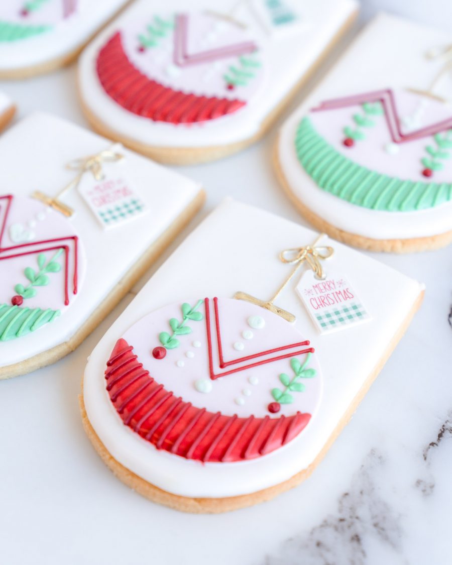 Hand-decorated festive ornament sugar cookies with intricate red and green patterns, golden accents, and mini holiday tags, arranged on a marble surface