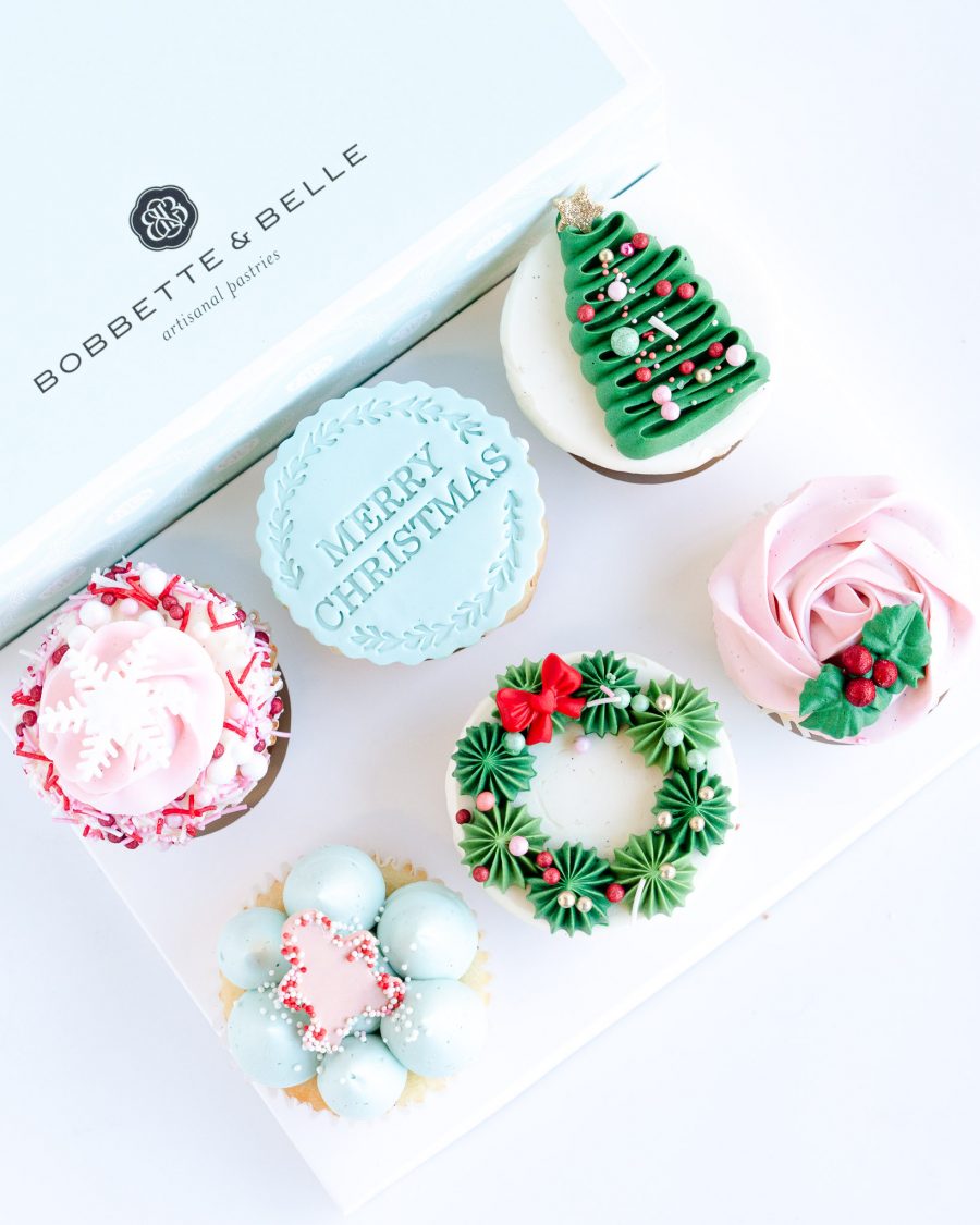 A 6-pack of holiday cupcakes with Christmas tree, wreath, and snowflake designs, topped with tinted vanilla buttercream