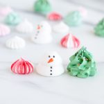 A clear round container filled with holiday-themed meringues, including snowman shapes, Christmas tree designs, and swirled red, green, and white meringue kisses.