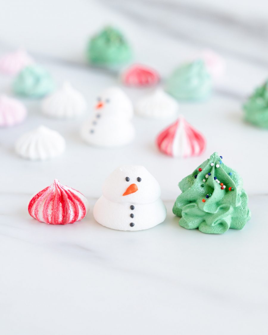 A clear round container filled with holiday-themed meringues, including snowman shapes, Christmas tree designs, and swirled red, green, and white meringue kisses.