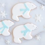 Adorable polar bear-shaped sugar cookies with white royal icing, sparkling blue scarves, and snowflake decorations, displayed on a marble surface.