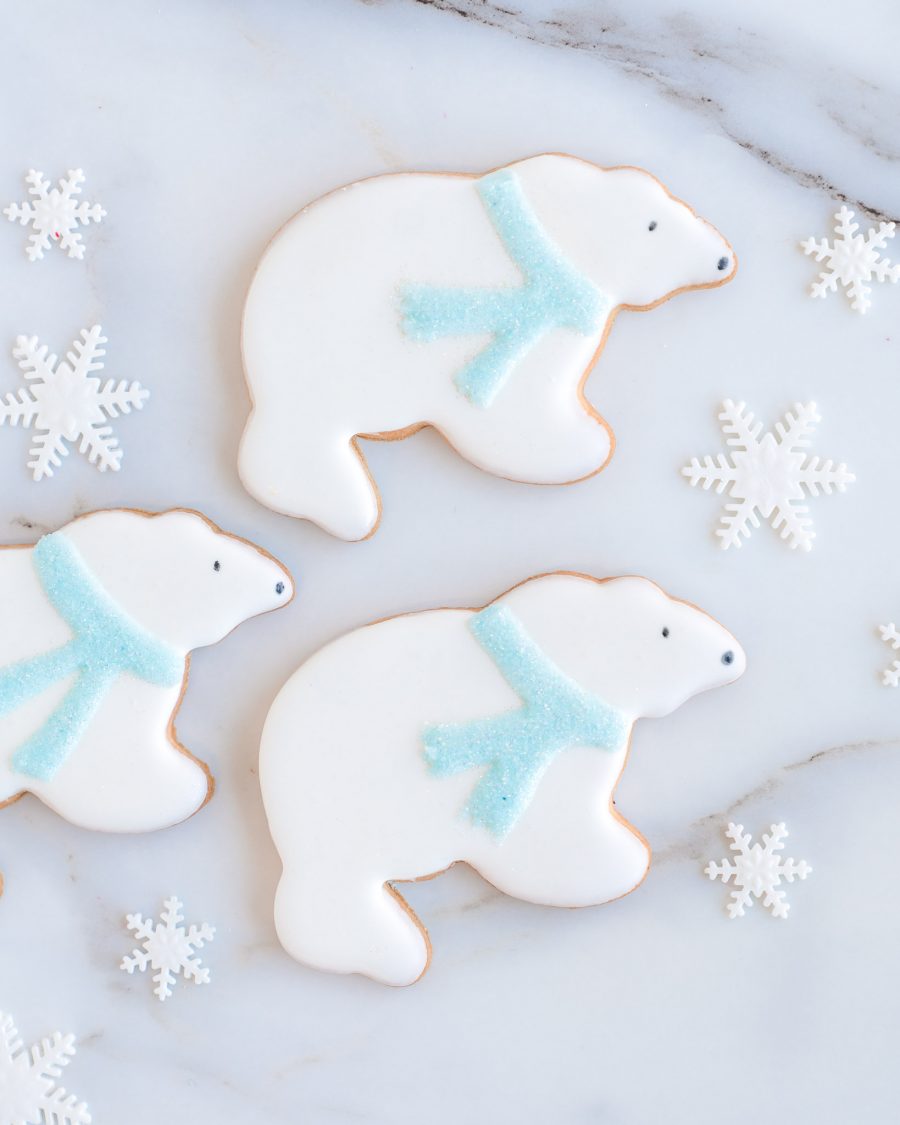 Adorable polar bear-shaped sugar cookies with white royal icing, sparkling blue scarves, and snowflake decorations, displayed on a marble surface.