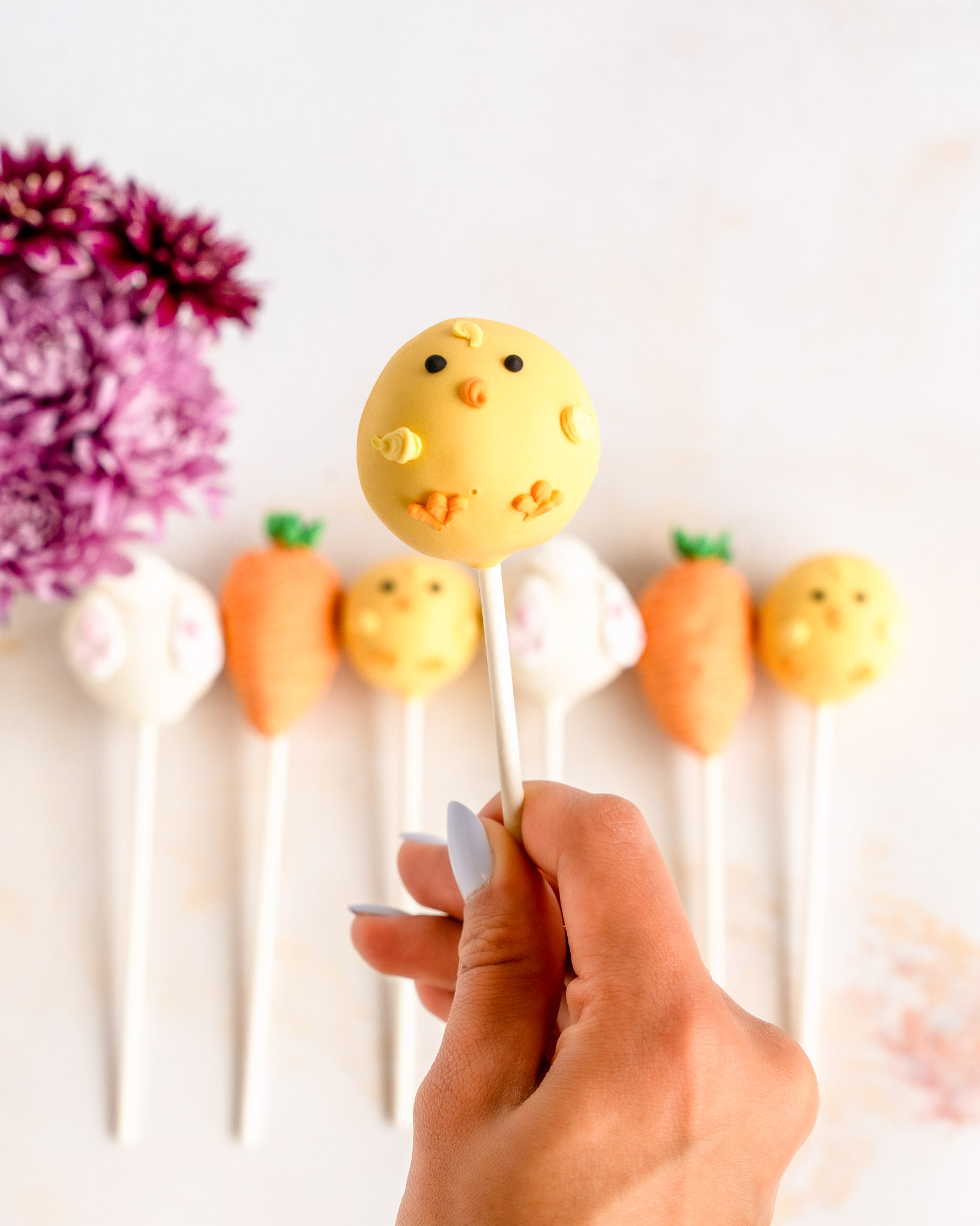 Hand holding Easter cake pops decorated as a chick, bunny tail, and carrot with pastel chocolate coating and detailed designs