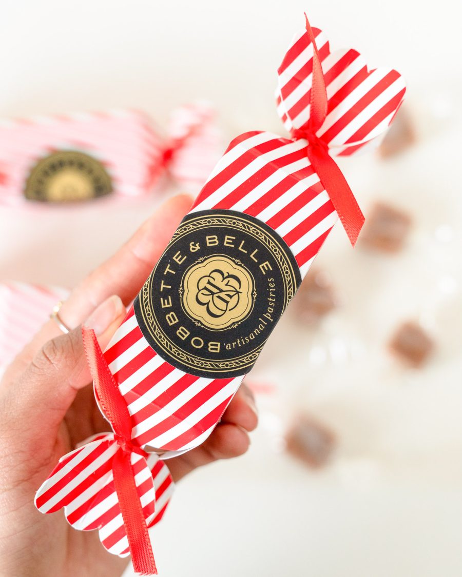 Salted Caramel Stocking Stuffer 2 Hand holding a red-and-white striped Bobbette & Belle holiday caramel cracker with gold and black artisanal label.
