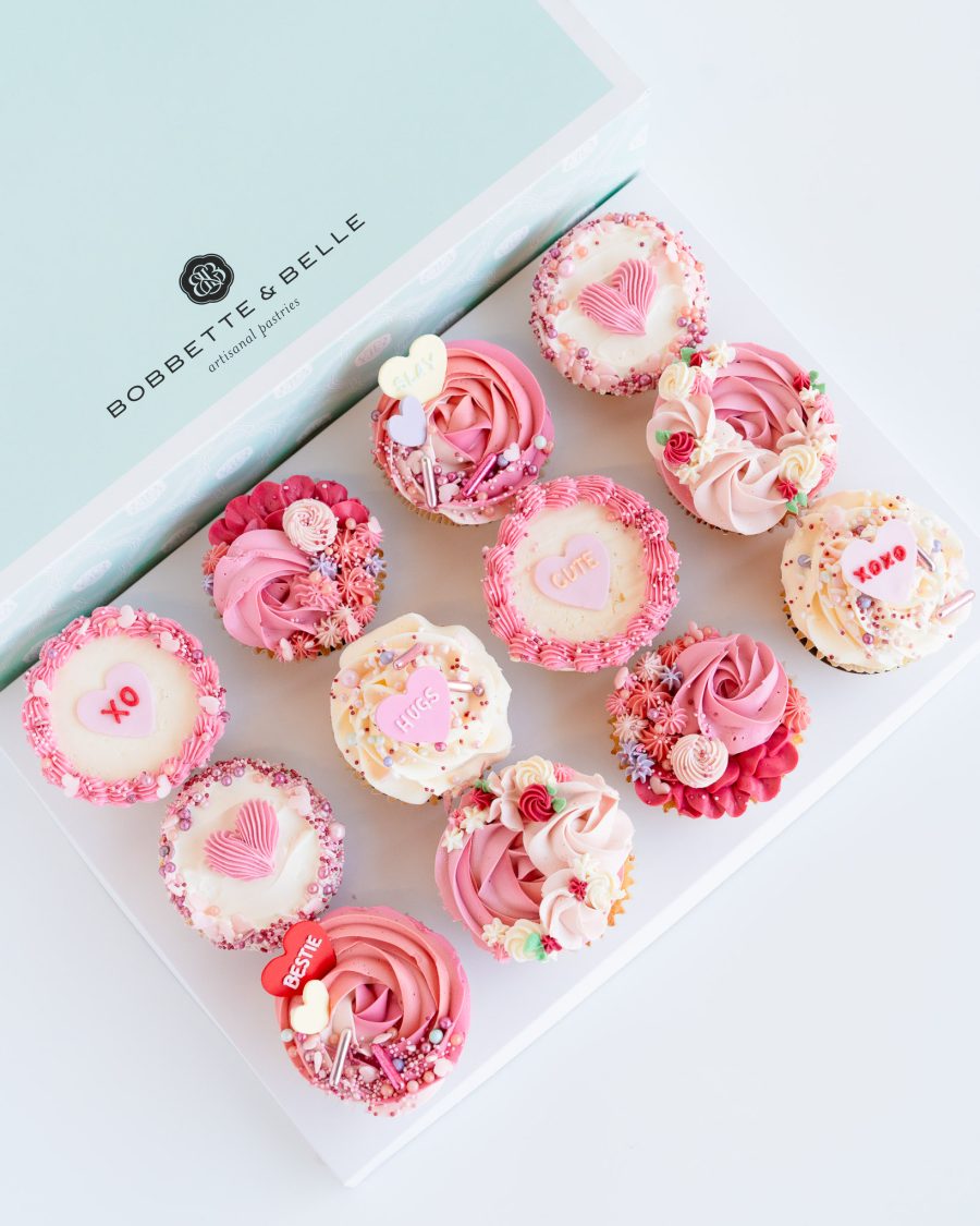 Box of 12 Valentine’s cupcakes decorated with tinted vanilla buttercream swirls and romantic heart toppers.
